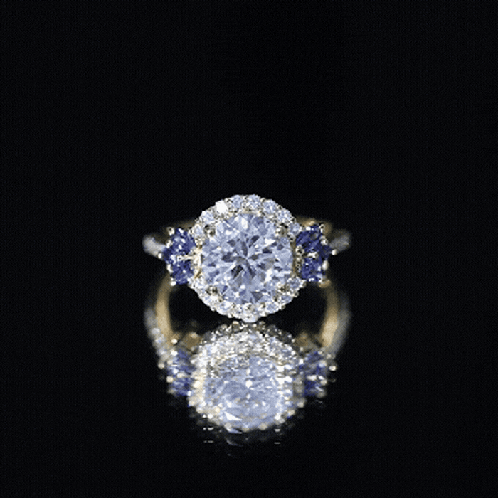 Yellow gold halo engagement ring with a 3-carat lab-grown diamond and blue sapphire accent, featuring intricate detailing.