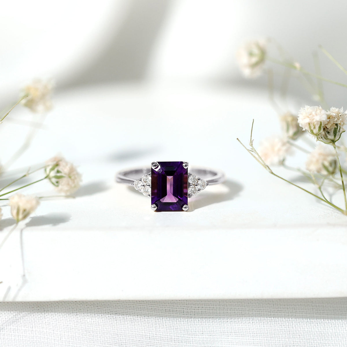 Emerald cut 1.4 carat amethyst engagement ring with diamond trio setting from Rosec Jewels, showcasing a silver band and vibrant purple gemstone.