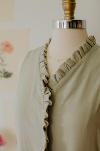A woman wearing The Village Green house dress with a relaxed fit, featuring a floral pattern and short sleeves, standing in a cozy room.